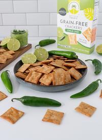 Artisan Tropic Chile Lime Crackers in a bowl by a board with guacamole, limes, and jalapeño peppers