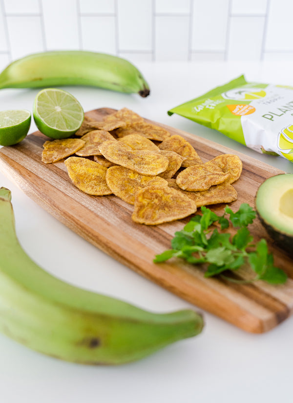 Cutting board with Artisan Tropic's Sea Salt Plantain Strips, limes, avocado and green plantains in the background